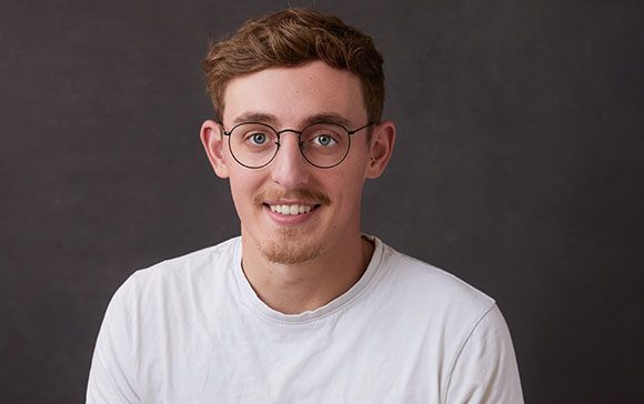 A young man with light brown hair and round glasses smiling in a white t-shirt.