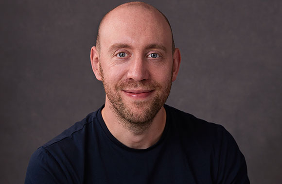 A smiling bald man with light eyes and a short beard wearing a dark t-shirt against a gray background.