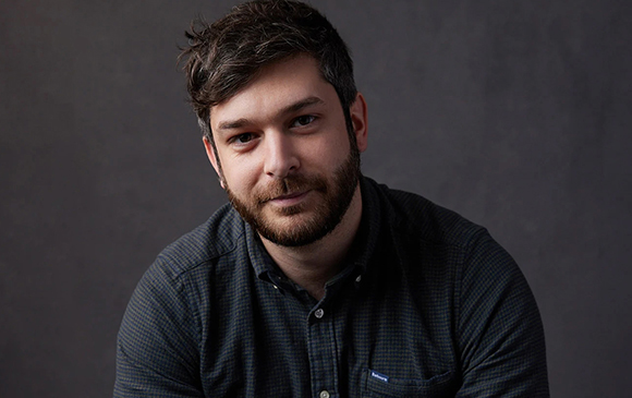 A man with dark hair and a beard wearing a dark patterned button-down shirt against a gray background.
