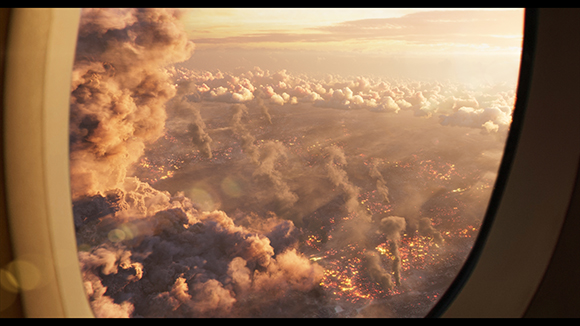 View from an airplane window showing large smoke plumes and glowing fires across a landscape at dusk.