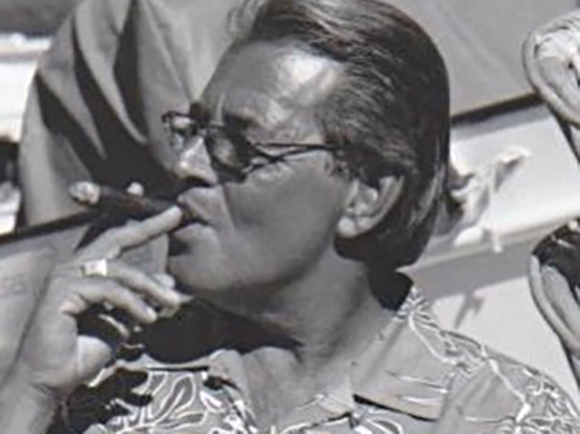 Black and white close-up of a man wearing glasses and a patterned shirt while smoking a cigar.