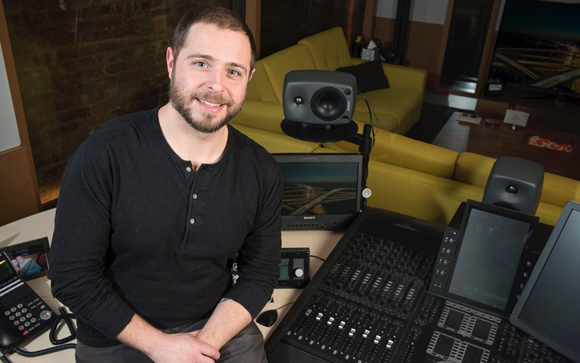 A man in a black shirt smiles while sitting in a studio with a mixing console and monitors.