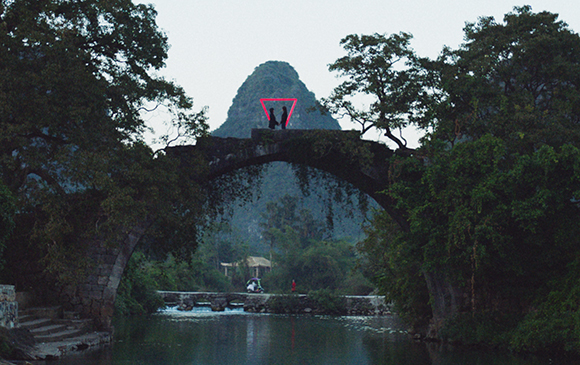 Two people stand on a stone arch bridge overlooking a river with a large mountain behind them.