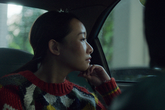 A woman in a colorful sweater looks out a car window with her hand near her chin.