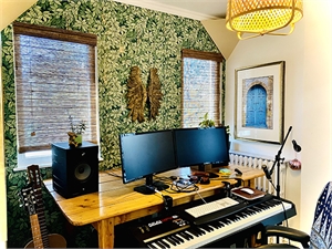 A home studio featuring a wooden desk with dual monitors, speakers, a keyboard, and a guitar against leafy wallpaper.