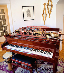 A dark wood upright piano with a decorative wooden screen, a djembe drum, and a small brass bowl.