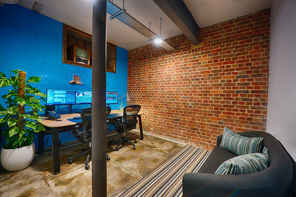 Office workspace with a desk, computer monitors, a potted plant, and a dark sofa against brick and blue walls.