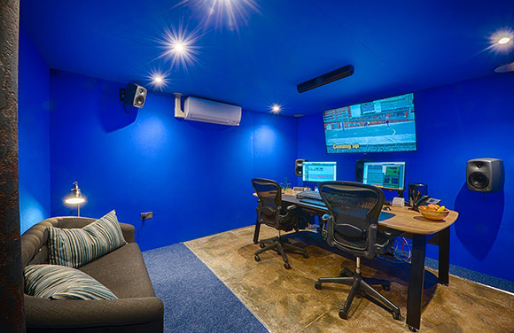 A blue media room featuring a desk with dual monitors, office chairs, a large wall-mounted screen, and a sofa.