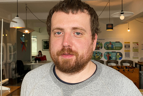 A man with short brown hair and a reddish-brown beard looking forward in an office.