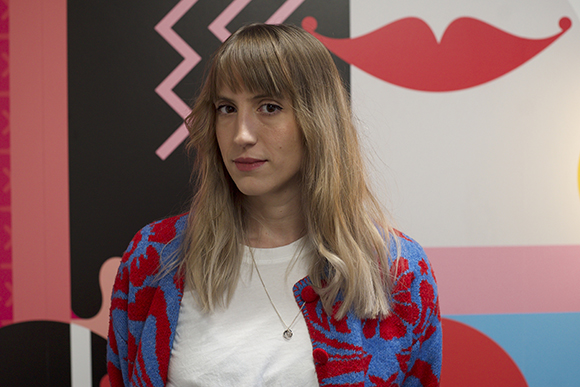A woman with long blonde hair and bangs wearing a blue and red patterned cardigan over a white shirt.