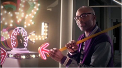 A man in glasses and a purple jacket holds a long yellow rod near glowing marquee lights.