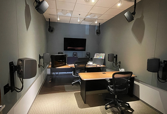 A small studio room with gray walls, multiple mounted speakers, two desks, and office chairs.
