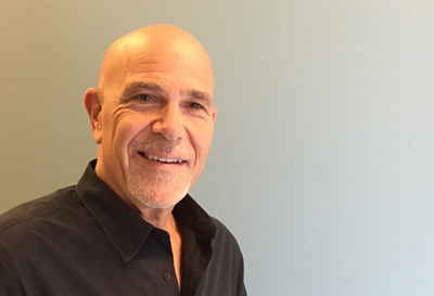 A smiling bald man with a graying beard wearing a black button-down shirt against a plain background.