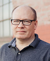 A man with short light hair and black-rimmed glasses wearing a grey polo shirt.