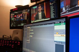 Multiple computer monitors in a control room displaying news broadcasts and technical production software.