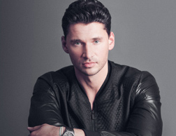 A man with dark hair and blue eyes wearing a black textured leather jacket against a gray background.