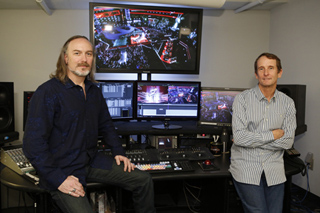 Two men stand in a studio filled with multiple computer monitors and video editing equipment.