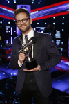 A smiling man with glasses in a navy suit holds a dark trophy on a stage with blue and red lights.