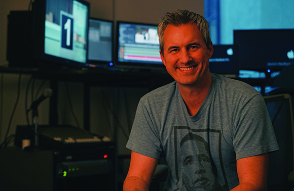A smiling man in a blue graphic t-shirt sits in a dark studio filled with computer monitors.