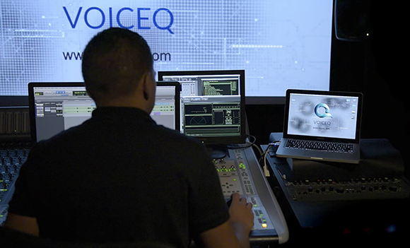 A person sits at a desk with three computer monitors and a mixing console in a dark studio.