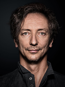 Close-up portrait of a man with dark, wavy hair and light eyes looking directly at the camera.