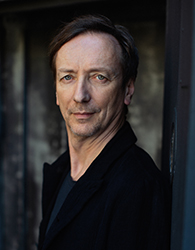 A man with short brown hair and light eyes wearing a black jacket looks toward the camera.