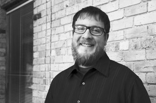 A smiling man with glasses and a beard wearing a dark button-down shirt against a brick wall.