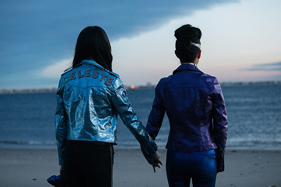 Two women seen from behind wearing shiny blue and purple jackets, standing on a beach at dusk.
