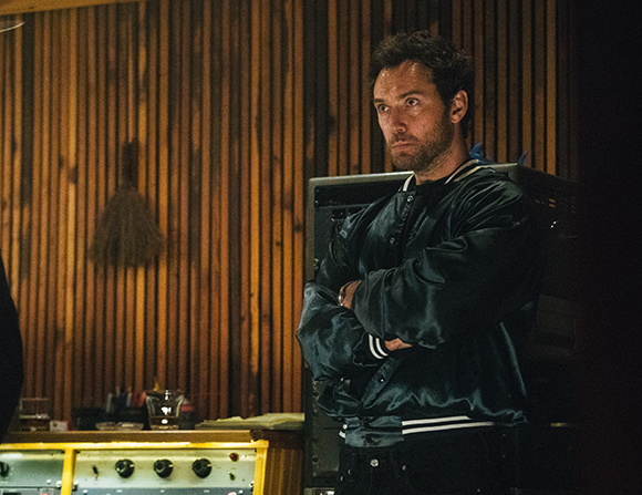A man with short dark hair and stubble stands with arms crossed wearing a dark satin bomber jacket.