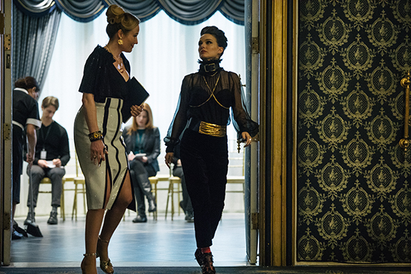 Two women in elegant evening wear walk through a doorway in a grand, decorated room.