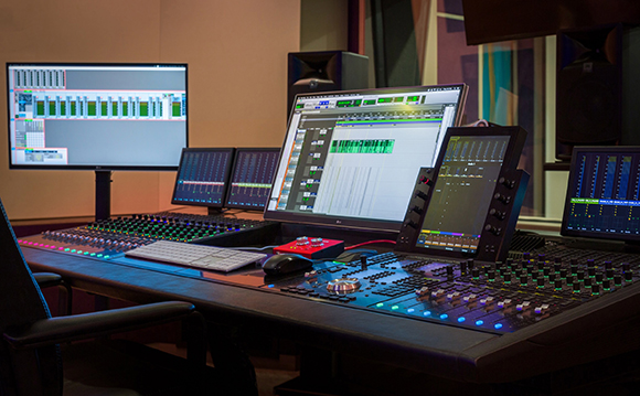 Professional audio mixing desk with multiple computer monitors and studio speakers in a dark studio.