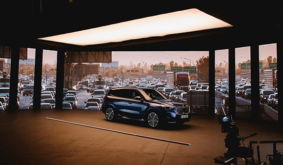 A blue SUV parked in a dark studio in front of a large screen displaying a sunset highway traffic jam.