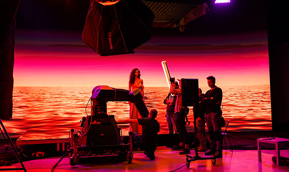 Film crew using cameras and lights in front of a large digital screen displaying a red sunset over water.