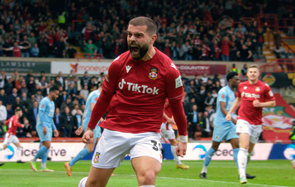 A male soccer player in a red jersey and white shorts shouts in celebration on a field.
