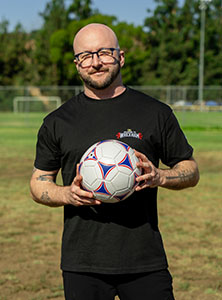 A bald man with glasses and a beard holds a white, red, and blue soccer ball in an outdoor field.