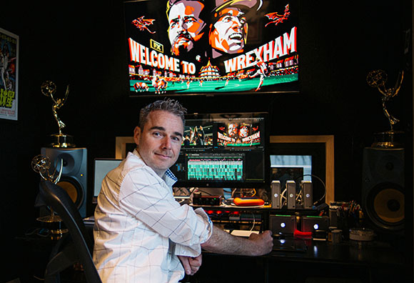A man sits in a dark studio facing the camera, with a large screen behind him displaying 'Welcome to Wrexham'.