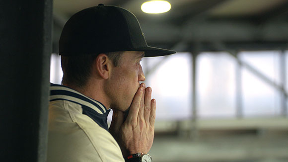Profile of a man wearing a black baseball cap and tan jacket, resting his chin on his hands.