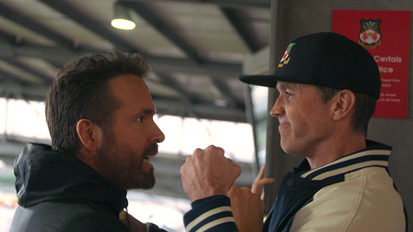 Two men in profile looking at each other, one wearing a dark jacket and the other a baseball cap and varsity jacket.