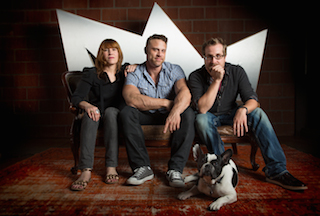 Three adults sit on a couch with a small black and white dog on a patterned rug in front of a white crown.