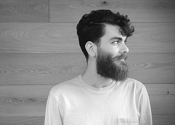 Black and white profile portrait of a man with dark wavy hair and a full beard against a wooden wall.