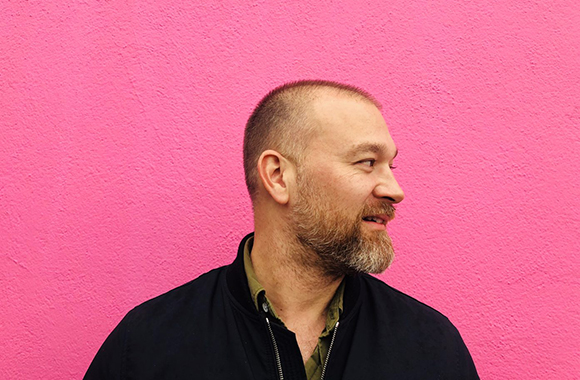 A bearded man in a black jacket looking to his right against a bright pink textured wall.
