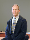 A man with light hair wearing a blue jacket, white shirt, and blue patterned tie against a gray background.