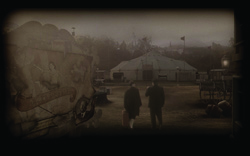 Sepia view of a circus camp with a large tent, wagons, and two people walking in a dark field.