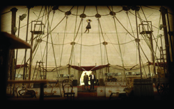 A tightrope walker balances on a wire high above a circus ring inside a large tent.