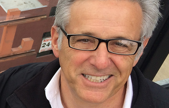 A smiling man with short gray hair and black-rimmed glasses wearing a white shirt and black jacket.