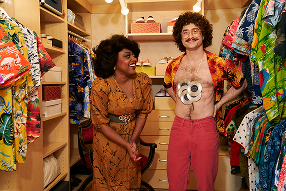 A woman in a brown dress and a man in an open patterned shirt and red pants smile in a clothing closet.