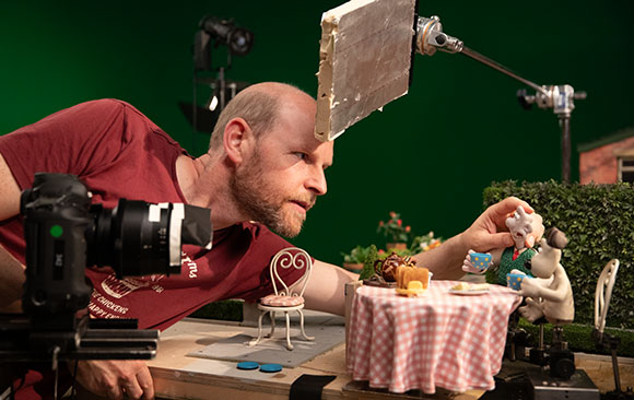 A man leans over a miniature table set with small food items and figurines during a film shoot.