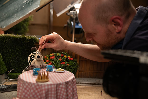 A man leans in to adjust a small clay dog figure on a miniature table set with tiny tea cups.