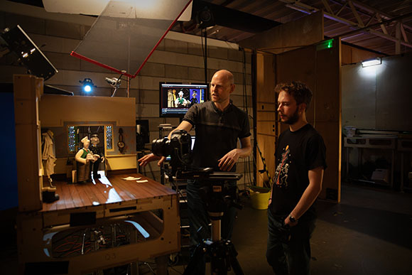 Two men stand in a studio near a miniature set and a professional camera on a tripod.