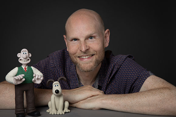 A smiling man leans on a table next to a clay man and a clay dog.